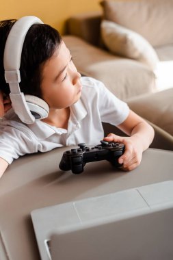 KYIV, UKRAINE - 22 Nisan 2020: Hüzünlü Asyalı çocuk joystick ve dizüstü bilgisayarla oyun oynuyor
