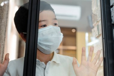 asian boy in medical mask looking through window on quarantine clipart