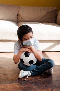 Futbol topuyla evde karantinada oturan üzgün Asyalı çocuk. 