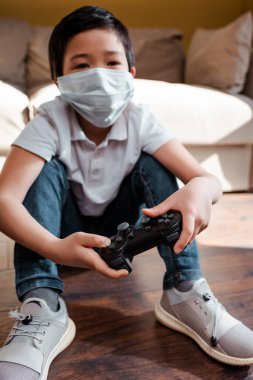 KYIV, UKRAINE - 22 Nisan 2020: tıbbi maskeli Asyalı çocuk karantinada joystick ile video oyunu oynuyor