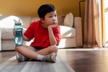 Biberonlu sıkılmış Asyalı çocuk evde spor minderinin üzerinde tek başına oturuyor.