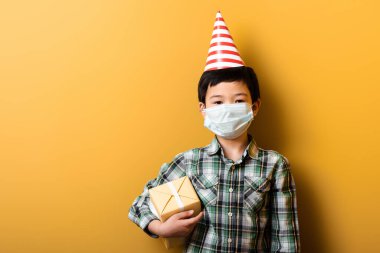 asian boy in party cone and medical mask holding birthday gift on yellow during self isolation clipart