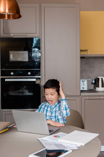 эмоциональный мальчик кричит с поднятой рукой и учится онлайн с ноутбуком дома во время самоизоляции