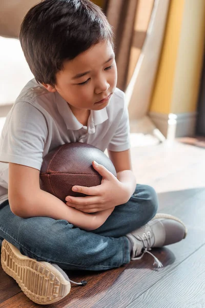 sad asian boy with rugby ball sitting on floor at home on self isolation