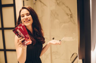 Siyah elbiseli gülümseyen kız dudak parlatıcısı tutuyor ve gece partisi biletleri banyoda, karantina konseptinin sonu. 