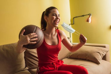 Heyecanlı sporcu kadın tıbbi maskeye dokunuyor ve basketbol topunu oturma odasında tutuyor, karantina konseptinin sonu. 