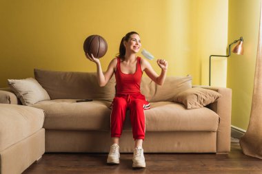Mutlu sporcu kadın tıbbi maskeye dokunuyor ve basketbol topunu oturma odasında tutuyor, karantina konseptinin sonu. 