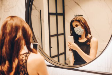 Siyah elbiseli ve tıbbi maskeli bir kadının banyoda fotoğraf çektiği seçici odak noktası.
