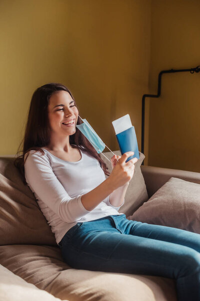 cheerful girl in medical mask holding passport with air ticket while sitting on sofa, end of quarantine concept