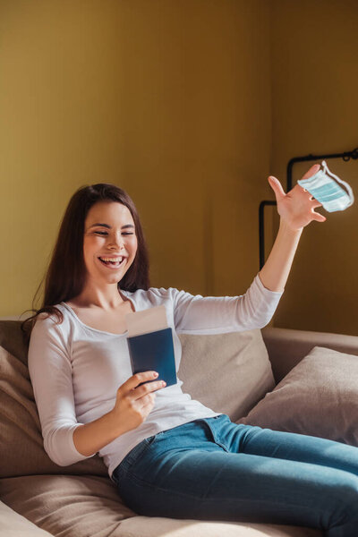 cheerful girl throwing in air medical mask and holding passport with air ticket while sitting on sofa, end of quarantine concept