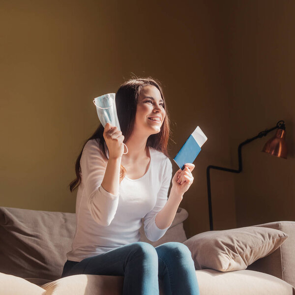 cheerful young woman holding passport with air ticket and medical mask while sitting on sofa, end of quarantine concept