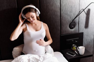 neşeli ve hamile bir kadın yatak odasında müzik dinlerken kulaklıklara dokunuyor. 