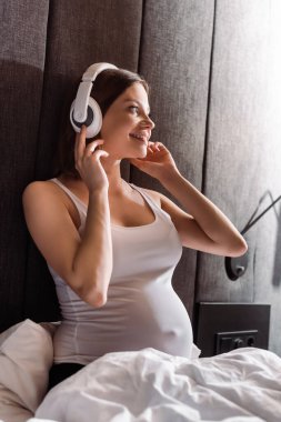 Mutlu ve hamile bir kadının yatak odasında müzik dinlerken kablosuz kulaklıklara dokunmasının seçici odağı 