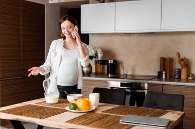 Mutlu ve hamile bir kadın mutfaktaki yemeğin yanında akıllı telefonla konuşurken el kol hareketi yapıyor. 