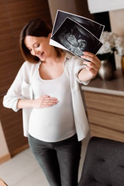 Ultrason fotoğrafları tutan neşeli ve hamile bir kadının seçici odağı 