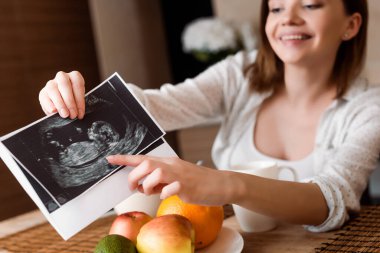 Seçici odak noktası, meyvelerin yanındaki ultrason fotoğraflarına parmak gösteren neşeli hamile kadın. 