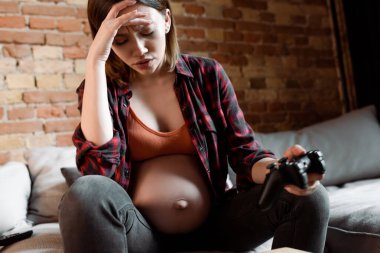 KYIV, UKRAINE - 30 Nisan 2020: oturma odasında elinde joystick tutan üzgün ve hamile bir kadının seçici odak noktası 