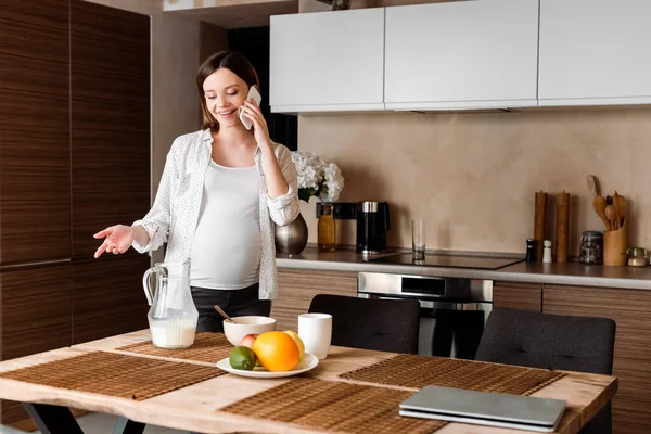 在厨房里 一个快乐而又怀孕的女人在智能手机边与食物交谈时做手势 — 图库照片