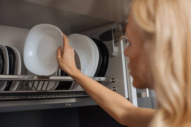 selective focus of young housewife putting clean plates on rack in kitchen clipart