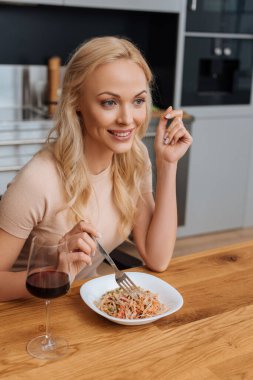 cheerful woman holding fork near plate with thai noodles and glass of red wine clipart