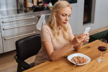 Genç bir kadın, Thai eriştesi ve bir bardak kırmızı şarapla tabağın yanında otururken akıllı telefondan sohbet ediyor.