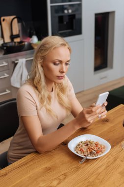 Ciddi bir kadın Thai eriştesiyle tabağın yanında otururken akıllı telefondan sohbet ediyor. 