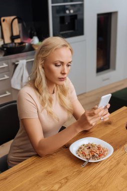 serious woman chatting on smartphone while sitting near plate with thai noodles  clipart