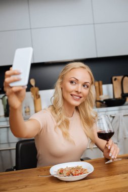 seçici odak noktası, gülümseyen bir kadının akıllı telefonuyla Thai erişteli kırmızı şarap ve tabağın yanında selfie çekmesi.