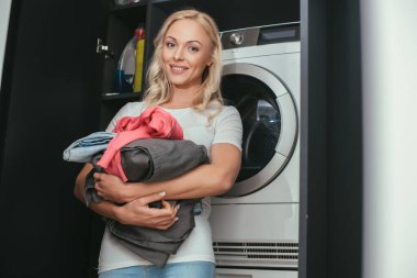 selective focus of smiling housewife looking at camera while holding laundry near washing machine clipart
