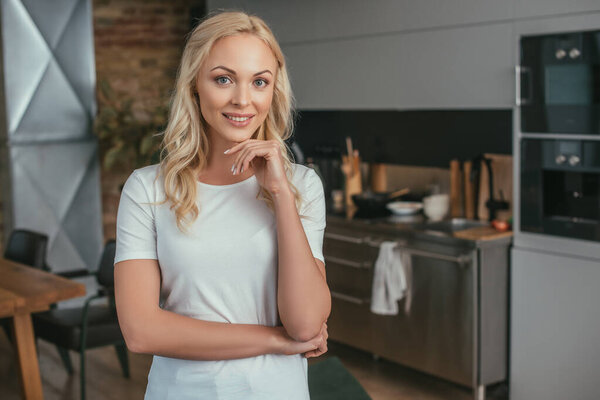 веселая блондинка смотрит в камеру и держит руку у подбородка, стоя дома
