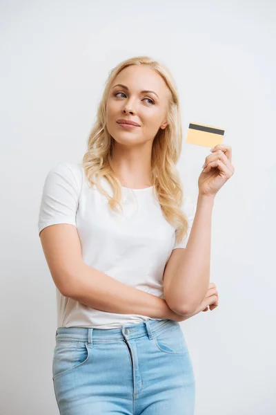 smiling, dreamy woman showing credit card while looking away isolated on white