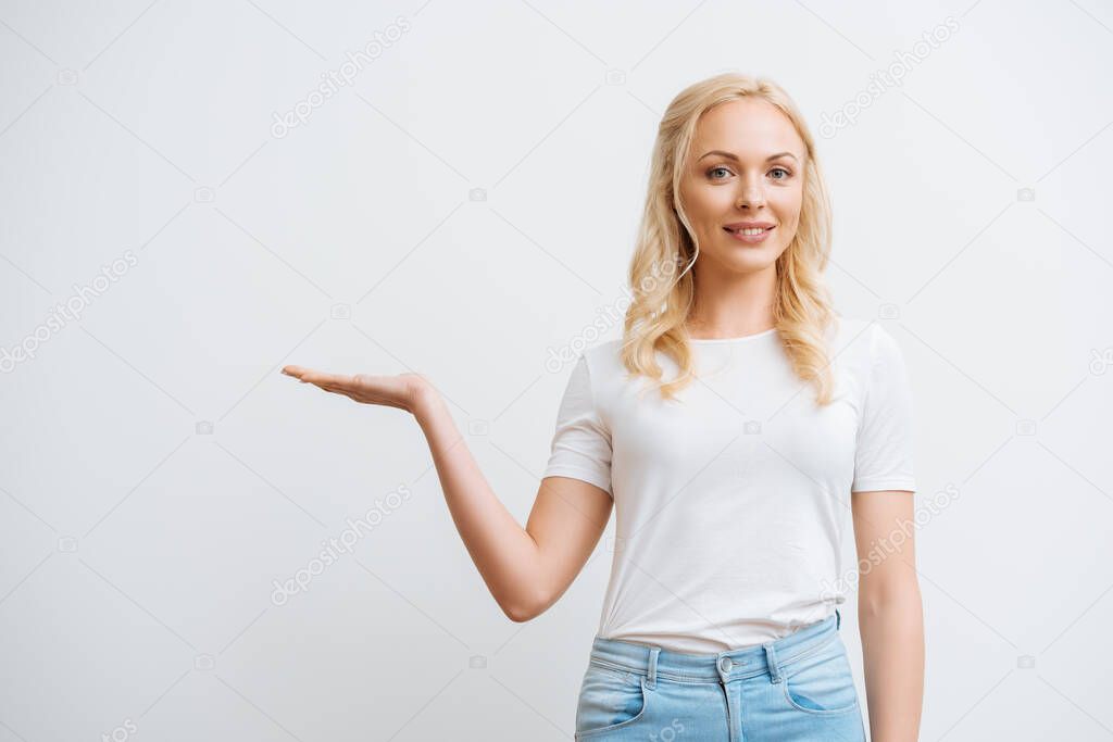 smiling blonde woman standing with open arm and looking at camera isolated on white