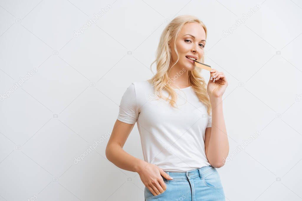 smiling woman holding credit card near lips while looking at camera isolated on white