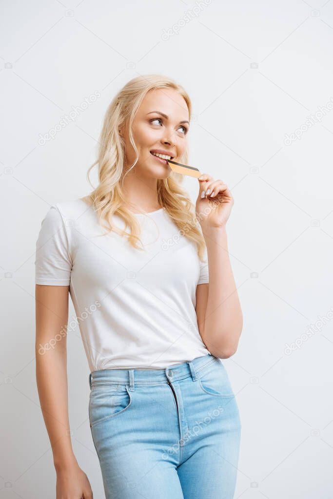 smiling, dreamy woman holding credit card near lips while looking away isolated on white