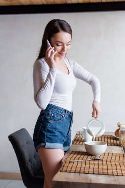 Mutlu kadın sürahiyi tutarken kaseye süt koyuyor ve akıllı telefondan konuşuyor. 