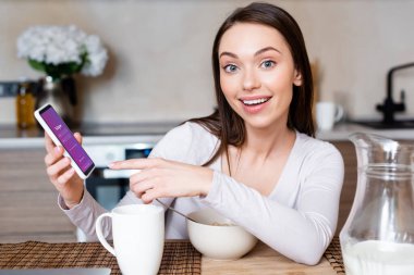 KYIV, UKRAINE - 29 Nisan 2020: mutlu bir kızın Instagram uygulaması ile akıllı telefonu işaret ederek kupa, kase ve sütle sürahiyi işaret etmesi 