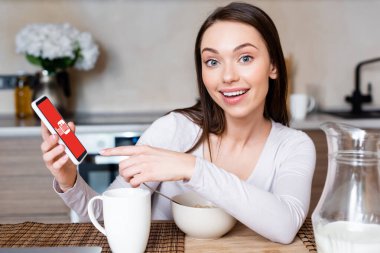 KYIV, UKRAINE - 29 Nisan 2020: akıllı telefonu bardağa yakın youtube uygulaması ile işaret eden mutlu kızın seçici odağı, kase ve sütle dolu sürahi 