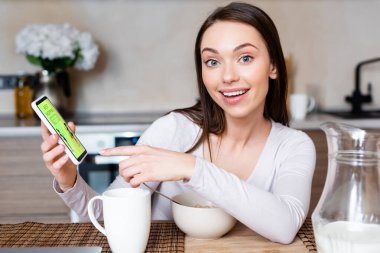 Akıllı telefonu işaret eden mutlu bir kızın seçici odak noktası. Yanında internet alışveriş uygulaması, kase ve sütle birlikte sürahi var. 