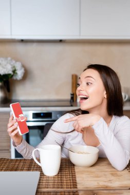 KYIV, UKRAINE - 29 Nisan 2020: akıllı telefonu işaret eden heyecanlı kız, Youtube uygulaması ile bardağa ve bowlinge yakın 