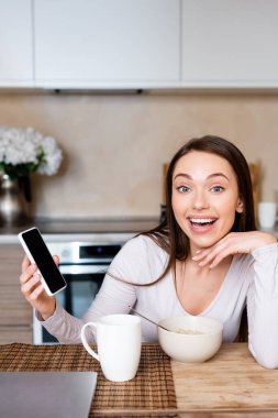 Heyecanlı kadın elinde boş ekranlı akıllı telefon, bardağın ve kasenin yanında. 