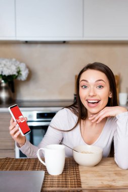 KYIV, UKRAINE - 29 Nisan 2020: Fincanın ve bowlingin yanında Youtube uygulaması ile akıllı telefon tutan heyecanlı kadın 