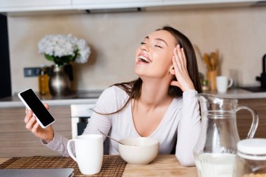 Mutlu kadın elinde boş ekranla akıllı telefon tutuyor ve kahvaltıya yakın gülüyor. 