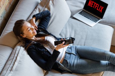 KYIV, UKRAINE - 29 Nisan 2020: Kanepe üzerinde netflix web sitesi olan dizüstü bilgisayarın yanında uzaktan kumandalı mutlu bir kadın görüntüsü 