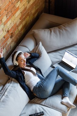 KYIV, UKRAINE - 29 Nisan 2020: Facebook sitesi olan dizüstü bilgisayarlı kablosuz kulaklıkla müzik dinleyen mutlu kadının genel görünümü 