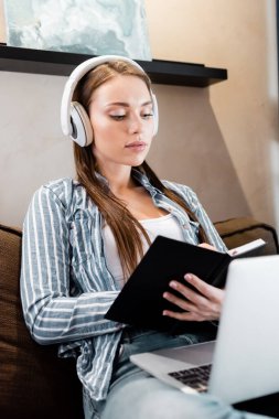 Kablosuz kulaklıklı çekici bir kadının oturma odasındaki dizüstü bilgisayarın yanında yazması, online çalışma konsepti. 