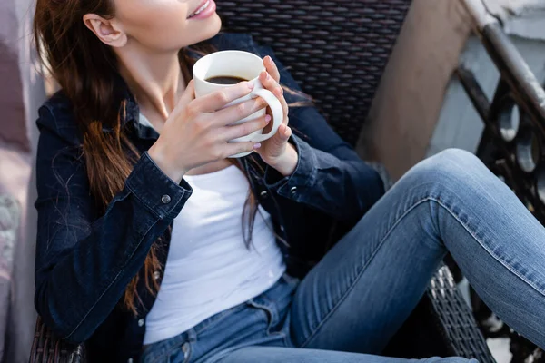 デニムジーンズやジャケットに身を包んだ幸せな女の子が椅子に座りバルコニーでコーヒーを飲みながら — ストック写真