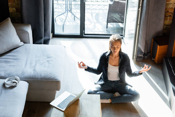 привлекательная молодая женщина с закрытыми глазами сидит в позе лотоса возле ноутбука
 