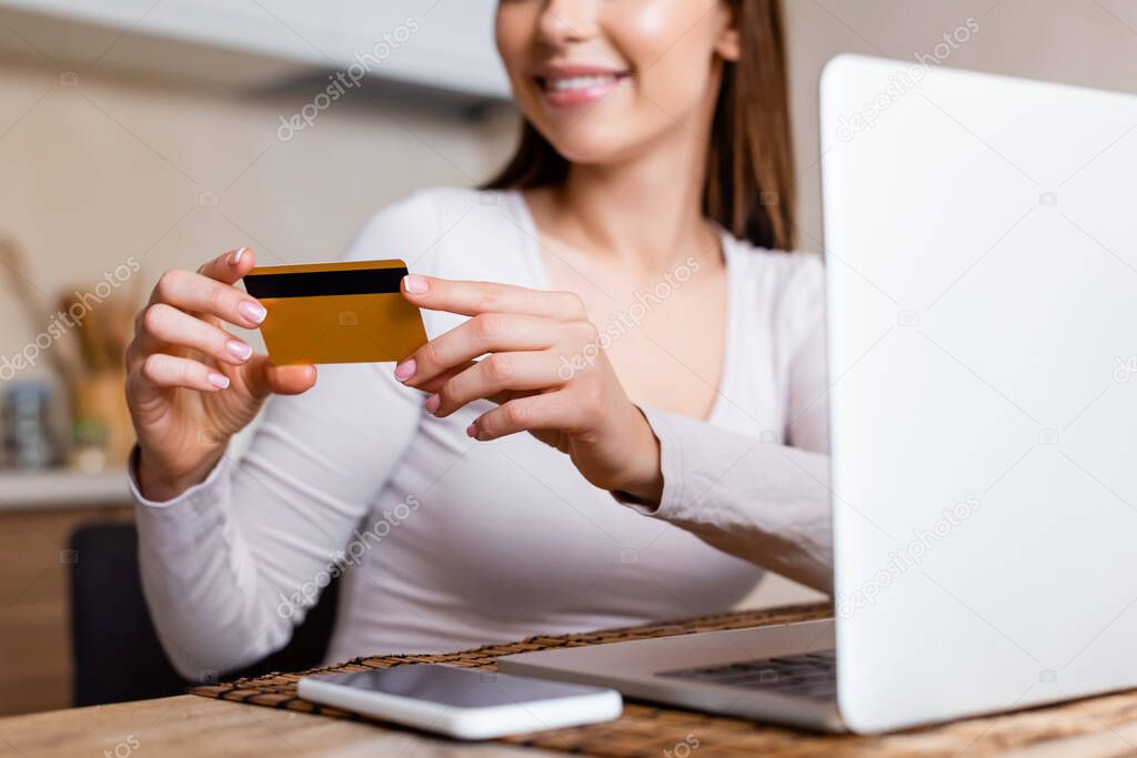 cropped view of happy girl holding credit card near laptop and smartphone on table 