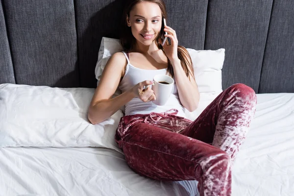 Leende Kvinna Talar Smartphone Och Hålla Kopp Kaffe Sängen — Stockfoto