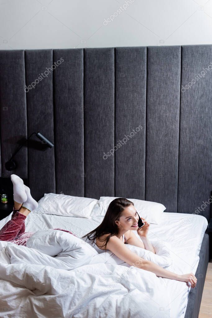 cheerful young woman talking on smartphone while lying on bed 